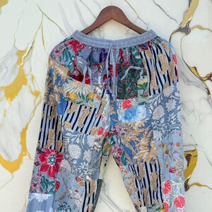 Puede incluir: Pantalones patchwork con cintura elástica gris y cordón. Los pantalones presentan una mezcla de estampados florales, a rayas y animales en tonos azules, rojos y blancos. Los pantalones están colgados de una percha de madera.