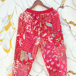 Puede incluir: Pantalones rojos estampados con cintura con cordón. Los pantalones presentan un diseño de patchwork con estampados florales y botánicos en tonos rojos, rosas y blancos. Los pantalones están colgados de una percha de madera.
