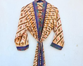 May include: A long, patterned robe with a tie closure, hanging on a wooden hanger. The robe features a repeating diamond pattern in shades of brown, tan, and red, with blue and patterned trim. The robe is made of a silky material.