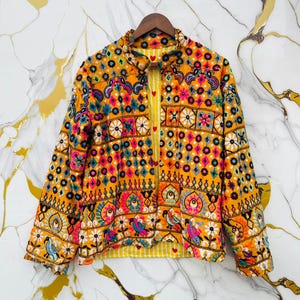 Könnte beinhalten: Eine bestickte Jacke in Gelb, Rosa, Blau und Orange. Die Jacke hat ein detailliertes Blumen- und geometrisches Muster mit einem gelb-weiß gestreiften Futter. Sie hängt an einem Holzkleiderbügel. Die Länge beträgt ca. 60 cm.
