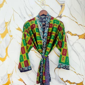 Op de afbeelding: Een kleurrijke kimono-badjas met een geometrisch patroon in groen, rood en geel. De badjas is omkeerbaar, met een bloemenpatroon aan de binnenkant. Hij wordt in de taille vastgebonden met een bijpassende sjerp. De badjas hangt aan een houten hanger.
