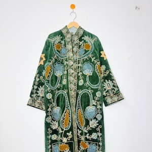 May include: A long-sleeved, knee-length coat made of dark green velvet fabric. The coat features intricate embroidery in shades of light blue, mustard yellow, and white, depicting floral and paisley-inspired patterns across the front.