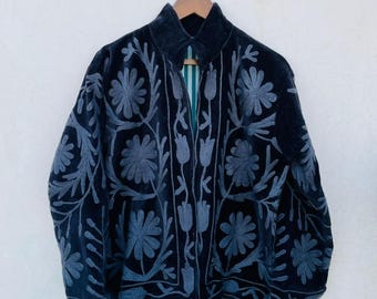 Pode incluir: Casaco azul marinho escuro com bordados florais cinzentos. O casaco está pendurado num cabide de madeira. Os bordados apresentam padrões detalhados de flores e folhas.