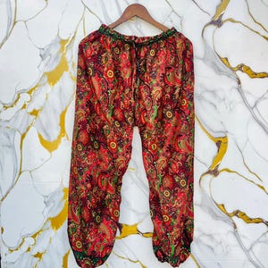May include: Red, paisley-patterned pants with a drawstring waist and elastic cuffs. The pants feature a vibrant mix of red, green, and yellow paisley designs. The pants are hanging on a wooden hanger.