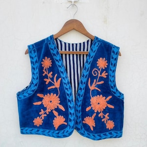 May include: A sleeveless vest in a rich blue colour, embellished with orange floral embroidery. The vest has a blue trim and a striped lining. The garment is displayed on a wooden hanger against a white background.