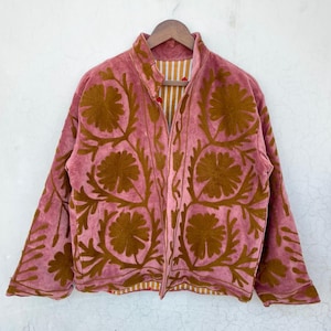 May include: A dusky pink jacket with brown floral embroidery. The jacket has a high collar and long sleeves. The interior lining features a striped pattern in shades of yellow and white. The jacket is hanging on a wooden hanger.