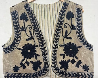May include: A beige vest with black floral embroidery and leaf patterns. The vest has a striped lining and is displayed on a wooden hanger. The embroidery creates a striking contrast against the beige fabric, showcasing a bohemian style.
