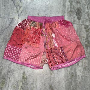May include: Pink patchwork shorts with a drawstring waist. The shorts feature a variety of pink fabric patterns, including floral, geometric, and dotted designs. The edges are trimmed with a solid pink fabric.