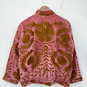 May include: A dusky pink jacket with a high collar and long sleeves. The jacket features detailed, embroidered floral patterns in a warm brown colour across the back and sleeves. The jacket is hanging on a wooden hanger.