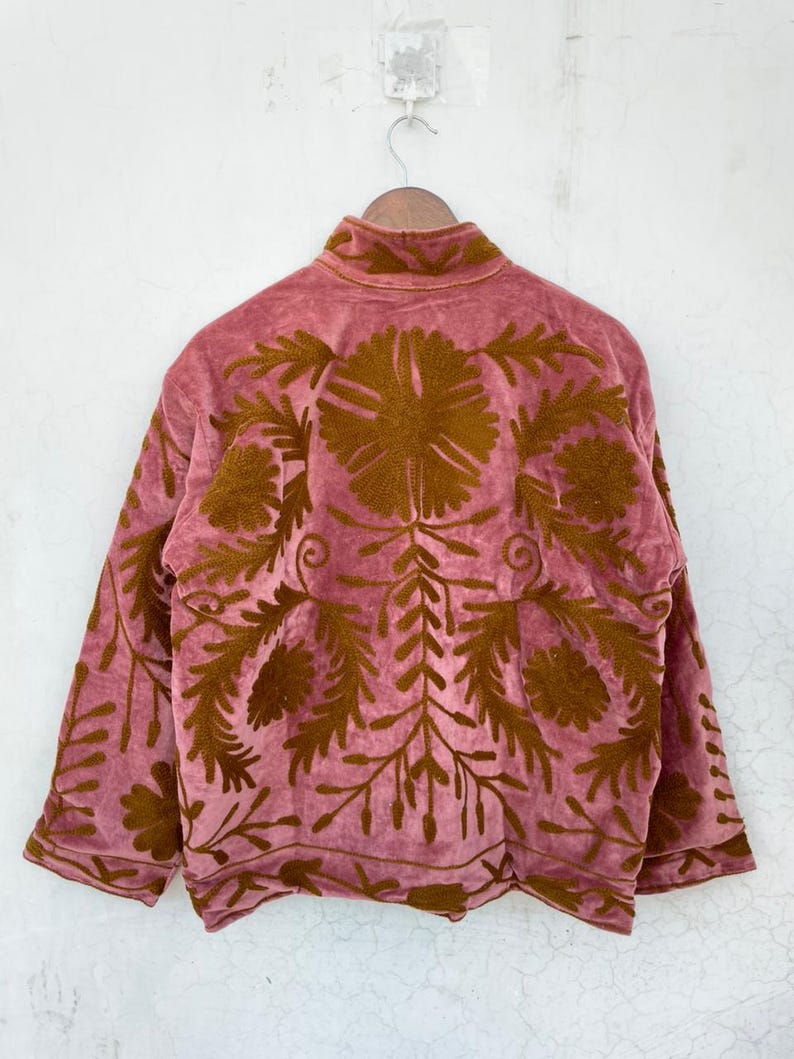 May include: A dusky pink jacket with a high collar and long sleeves. The jacket features detailed, embroidered floral patterns in a warm brown colour across the back and sleeves. The jacket is hanging on a wooden hanger.