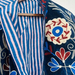 May include: A dark blue embroidered jacket with a striped blue and white lining. The jacket features floral embroidery in red, white, and blue. The jacket is hanging on a wooden hanger.