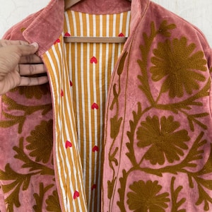 May include: A dusty rose jacket with brown floral embroidery. The jacket's interior features a striped pattern in gold and white, with small red heart accents. The jacket is hanging on a wooden hanger.