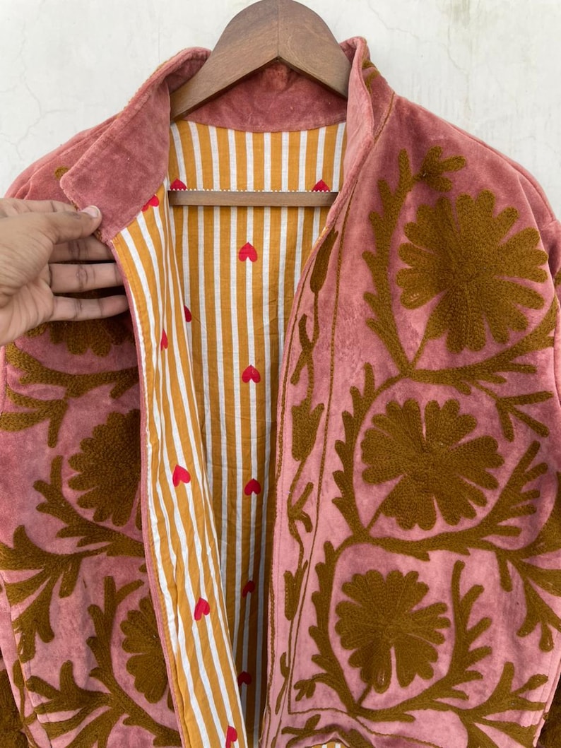May include: A dusty rose jacket with brown floral embroidery. The jacket's interior features a striped pattern in gold and white, with small red heart accents. The jacket is hanging on a wooden hanger.