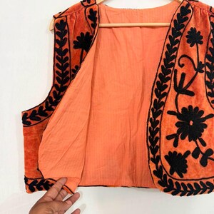 K&ouml;nnte beinhalten: Eine &auml;rmellose Weste in Orange mit schwarzen, gestickten Blumen- und Blattmustern entlang der Vorderkanten und des Saums. Das Kleidungsst&uuml;ck besteht aus einem weichen, samtartigen Stoff mit einem einfarbigen orangefarbenen Futter auf einem Holzkleiderb&uuml;gel.