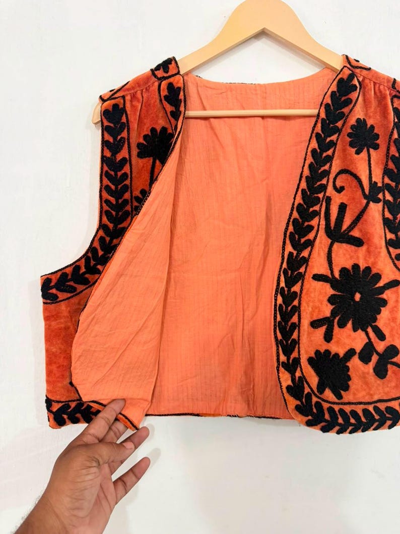 K&ouml;nnte beinhalten: Eine &auml;rmellose Weste in Orange mit schwarzen, gestickten Blumen- und Blattmustern entlang der Vorderkanten und des Saums. Das Kleidungsst&uuml;ck besteht aus einem weichen, samtartigen Stoff mit einem einfarbigen orangefarbenen Futter auf einem Holzkleiderb&uuml;gel.
