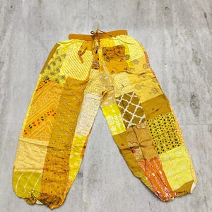 May include: Yellow patchwork pants with a drawstring waist. The pants feature a variety of patterns and shades of yellow, including floral, dotted, and geometric designs. The pants are made of a lightweight fabric.