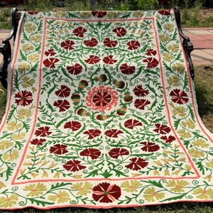 May include: Embroidered floral blanket in cream, red, green, and yellow. The blanket features a repeating pattern of red flowers with green leaves and stems, bordered by yellow and red floral designs. The blanket is draped over a black bench.