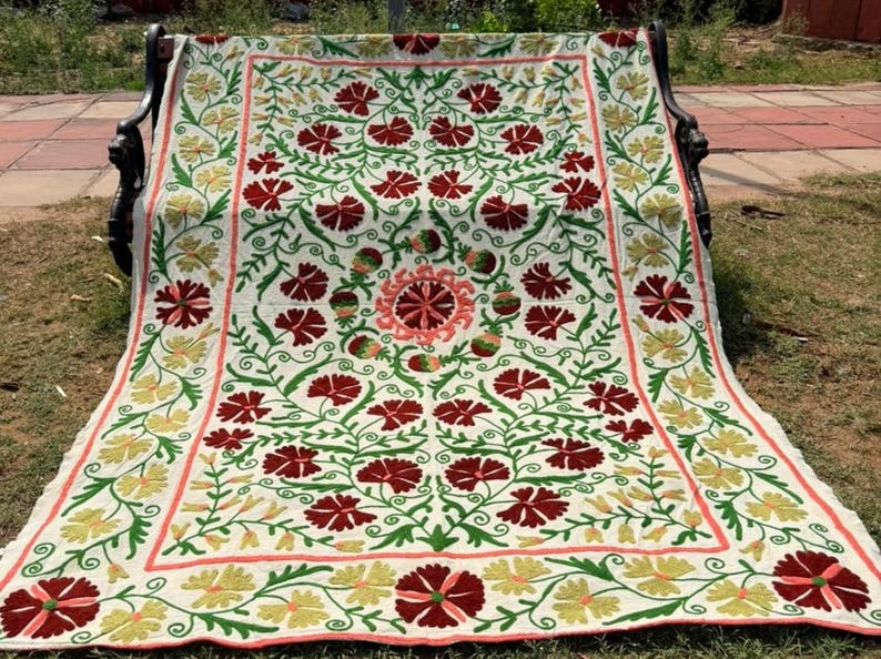 May include: Embroidered floral blanket in cream, red, green, and yellow. The blanket features a repeating pattern of red flowers with green leaves and stems, bordered by yellow and red floral designs. The blanket is draped over a black bench.