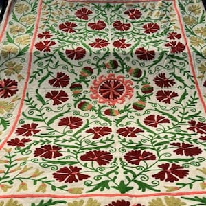 May include: A white textile with an intricate floral embroidery design. The embroidery features red flowers, green vines, and accents of yellow and pink. The pattern is symmetrical and covers the entire surface.