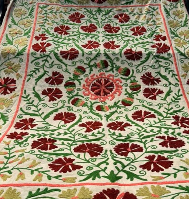 May include: A white textile with an intricate floral embroidery design. The embroidery features red flowers, green vines, and accents of yellow and pink. The pattern is symmetrical and covers the entire surface.