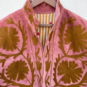 May include: A pink jacket with brown floral embroidery. The jacket has a high collar and a striped lining in shades of white and gold. The jacket is hanging on a wooden hanger.