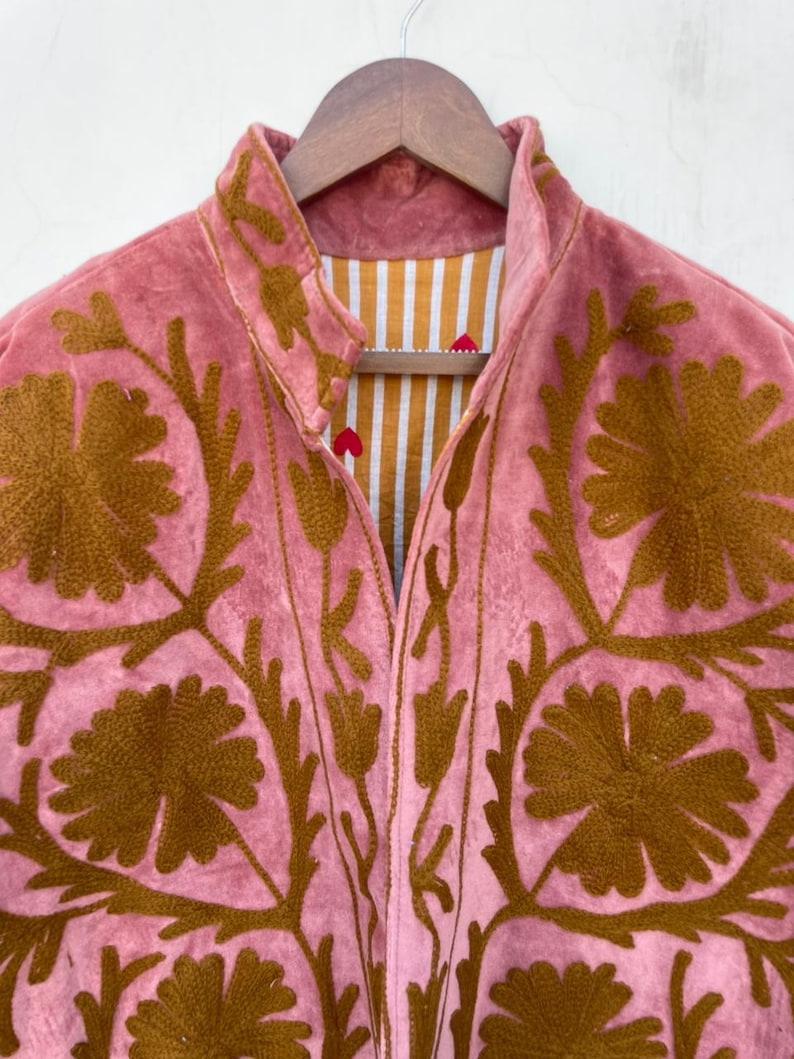 May include: A pink jacket with brown floral embroidery. The jacket has a high collar and a striped lining in shades of white and gold. The jacket is hanging on a wooden hanger.