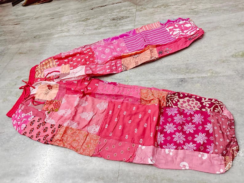 May include: Pink patchwork trousers with a drawstring waist. The trousers display a variety of pink and red patterns, including floral and geometric designs. The fabric appears lightweight and suitable for warm weather.