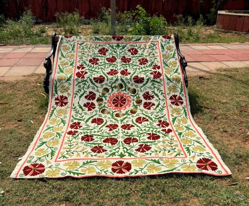 May include: A cream-colored, rectangular textile with a floral pattern. The design features red flowers, green leaves, and a border with yellow and pink accents. The textile is draped over a bench.