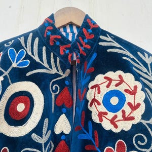 May include: A dark blue jacket with intricate floral embroidery in red, white, and blue. The jacket features a high collar and a striped lining. The embroidery includes stylized flowers, leaves, and geometric shapes, creating a detailed and artistic design.