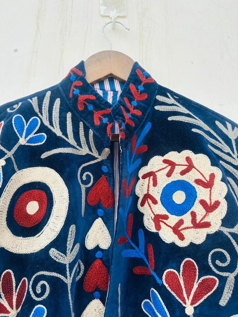 May include: A dark blue jacket with intricate floral embroidery in red, white, and blue. The jacket features a high collar and a striped lining. The embroidery includes stylized flowers, leaves, and geometric shapes, creating a detailed and artistic design.