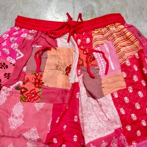 May include: Patchwork trousers with a red drawstring waist. The trousers are made from various pink and red fabrics, featuring floral and geometric patterns. The patchwork design creates a unique and colourful appearance.