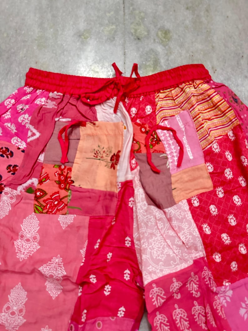 May include: Patchwork trousers with a red drawstring waist. The trousers are made from various pink and red fabrics, featuring floral and geometric patterns. The patchwork design creates a unique and colourful appearance.