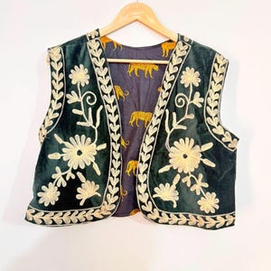 May include: A dark green velvet waistcoat featuring cream-coloured floral embroidery along the front edges and hem. The interior lining is a dark grey fabric decorated with a repeating pattern of yellow and black cheetahs, displayed on a wooden hanger.