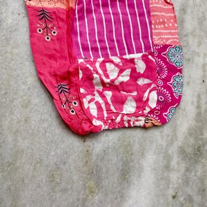 May include: A patchwork textile in shades of pink and white, featuring diverse patterns. The design incorporates vertical stripes, floral elements, and a pocket with a white and pink abstract pattern. The fabric is gathered at the base.