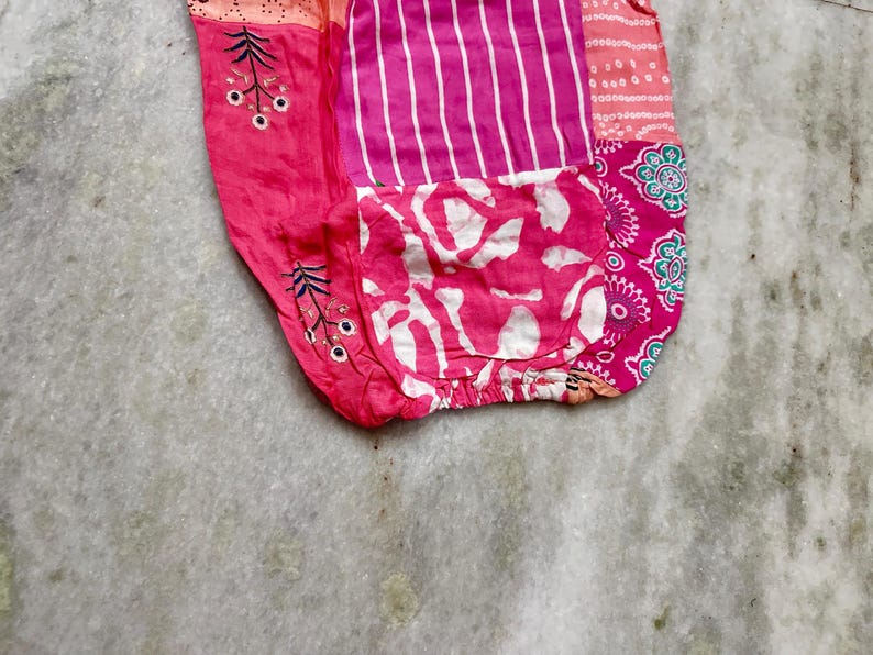 May include: A patchwork textile in shades of pink and white, featuring diverse patterns. The design incorporates vertical stripes, floral elements, and a pocket with a white and pink abstract pattern. The fabric is gathered at the base.