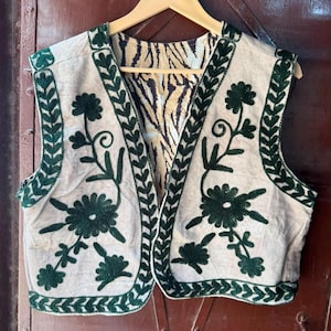 May include: A sleeveless waistcoat in a light beige fabric featuring dark green embroidered floral patterns along the edges and front panels. The interior lining displays a brown and white tiger stripe print. The garment is displayed on a wooden hanger.