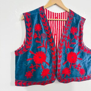 May include: A sleeveless vest featuring a deep blue velvet exterior with vibrant red floral embroidery along the front panels and edges. The interior lining is visible, showcasing a pattern of vertical red and white stripes throughout the garment.