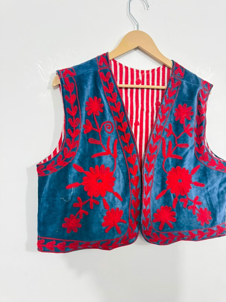 May include: A sleeveless vest featuring a deep blue velvet exterior with vibrant red floral embroidery along the front panels and edges. The interior lining is visible, showcasing a pattern of vertical red and white stripes throughout the garment.