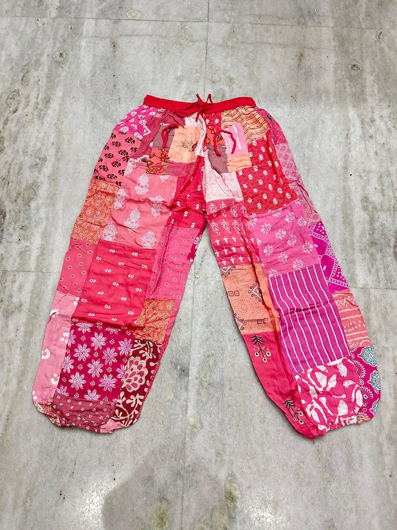 May include: Patchwork pants in shades of pink, red, and peach. The pants feature a drawstring waist and a variety of patterned fabric patches. The loose-fitting pants are laid flat on a light gray surface.