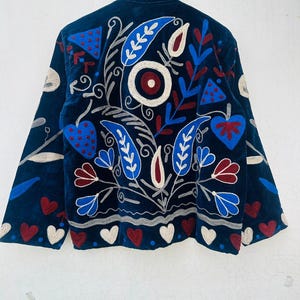 May include: A dark blue jacket with intricate embroidery. The design features red, white, and blue floral and heart motifs. The jacket has long sleeves and a high collar, with the embroidery covering the back and sleeves.