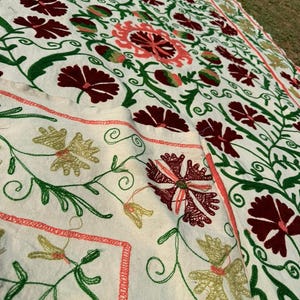 May include: Cream-colored textile with intricate floral embroidery. The design features burgundy, coral, and green flowers and foliage. The embroidery is detailed and covers the surface of the fabric, creating a textured appearance.