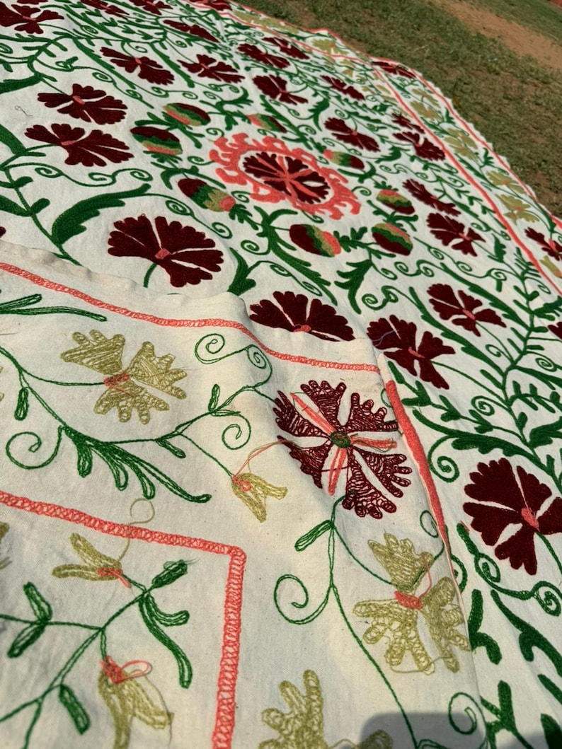 May include: Cream-colored textile with intricate floral embroidery. The design features burgundy, coral, and green flowers and foliage. The embroidery is detailed and covers the surface of the fabric, creating a textured appearance.