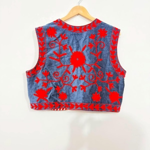 May include: A sleeveless vest in a muted blue fabric featuring vibrant red floral embroidery across the front. The design includes intricate vine and petal patterns along the neckline, armholes, and hem, with a red and white striped trim visible.