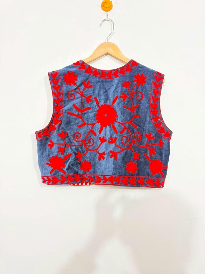 May include: A sleeveless vest in a muted blue fabric featuring vibrant red floral embroidery across the front. The design includes intricate vine and petal patterns along the neckline, armholes, and hem, with a red and white striped trim visible.