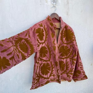 May include: A dusky pink jacket with a brown floral design. The jacket is suspended on a wooden hanger against a white wall. It features a zip-up front and a striped lining.