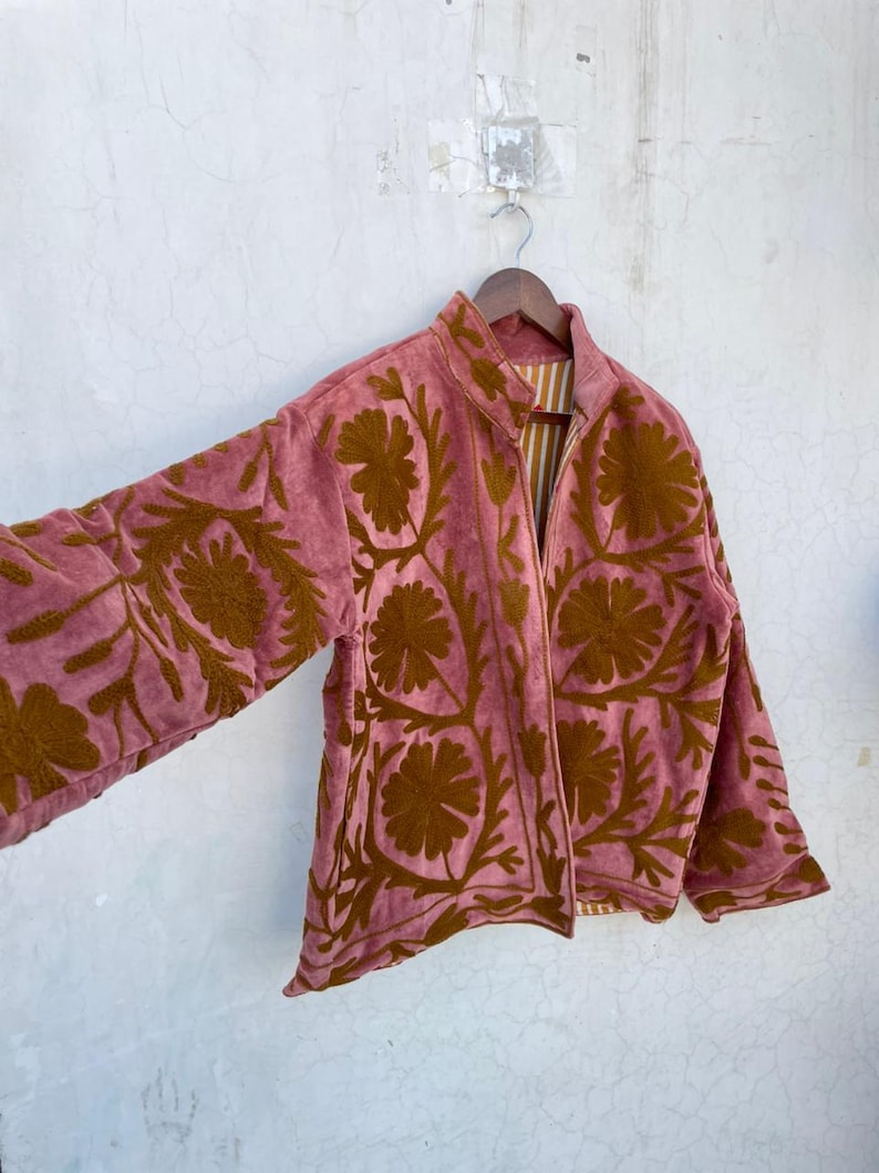 May include: A dusky pink jacket with a brown floral design. The jacket is suspended on a wooden hanger against a white wall. It features a zip-up front and a striped lining.