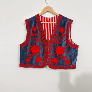 May include: A sleeveless vest featuring a dark blue velvet exterior with vibrant red floral embroidery along the edges and front panels. The interior lining displays a bold red and white vertical striped pattern, visible at the open front.