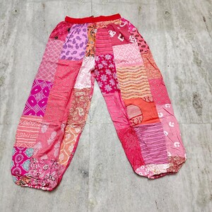 May include: Pink patchwork trousers with a red waistband. The trousers display a variety of pink and peach fabric patches with different patterns, including floral, geometric, and paisley designs. The trousers have a loose, comfortable fit.
