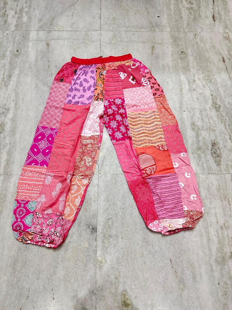 May include: Pink patchwork trousers with a red waistband. The trousers display a variety of pink and peach fabric patches with different patterns, including floral, geometric, and paisley designs. The trousers have a loose, comfortable fit.
