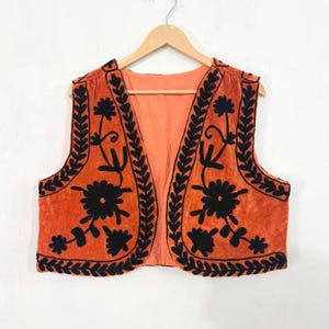 K&ouml;nnte beinhalten: Eine &auml;rmellose Weste in Orange mit schwarzer Blumen- und Blattstickerei entlang der Vorderkanten und des Saums. Das Kleidungsst&uuml;ck ist vorne offen gestaltet, hat ein hellorangefarbenes Innenfutter und h&auml;ngt an einem Holzkleiderb&uuml;gel.